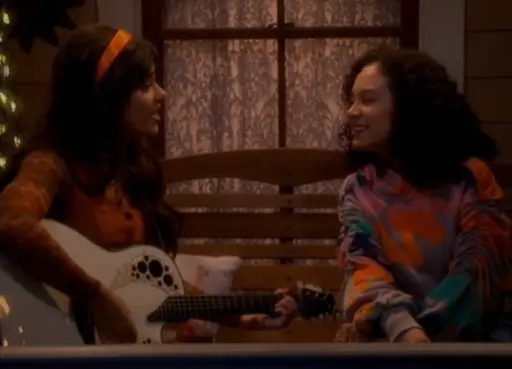 girl with an orange headband holding a guitar singing to a girl with dark curly hair.